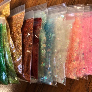 K&ouml;nnte beinhalten: Sortiment kleiner Beutel, gef&uuml;llt mit verschiedenen Farben von Glitter. Der Glitter ist schillernd und glitzert im Licht. Die Beutel sind aus durchsichtigem Plastik und in einer Reihe auf einer Holzoberfl&auml;che angeordnet.