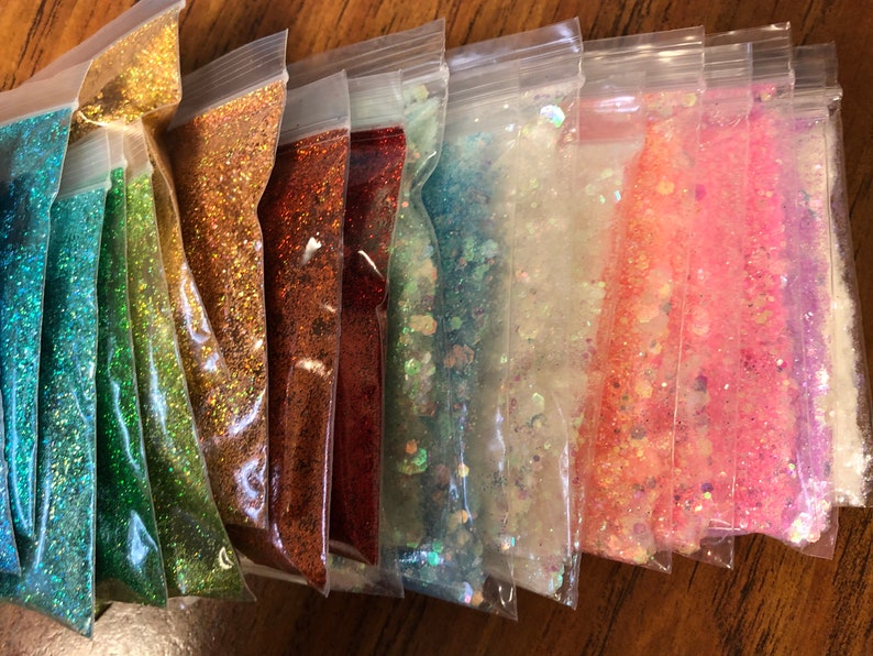 K&ouml;nnte beinhalten: Sortiment kleiner Beutel, gef&uuml;llt mit verschiedenen Farben von Glitter. Der Glitter ist schillernd und glitzert im Licht. Die Beutel sind aus durchsichtigem Plastik und in einer Reihe auf einer Holzoberfl&auml;che angeordnet.