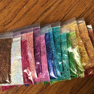 K&ouml;nnte beinhalten: Sortiment von 14 kleinen Beuteln mit schimmerndem Glitter in verschiedenen Farben, darunter Schwarz, Braun, Silber, Rosa, Lila, Blau, Gr&uuml;n, Gold und Rot.