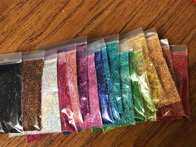 K&ouml;nnte beinhalten: Sortiment von 14 kleinen Beuteln mit schimmerndem Glitter in verschiedenen Farben, darunter Schwarz, Braun, Silber, Rosa, Lila, Blau, Gr&uuml;n, Gold und Rot.