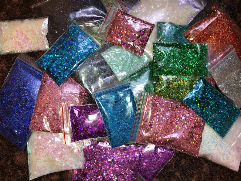 K&ouml;nnte beinhalten: Sortiment an Glitzer in verschiedenen Farben, darunter Blau, Gr&uuml;n, Pink, Lila und Wei&szlig;, verpackt in kleinen Plastikt&uuml;ten.