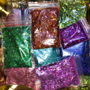 K&ouml;nnte beinhalten: Sortiment an schimmerndem Glitter in verschiedenen Farben, darunter Gr&uuml;n, Lila, Blau und Pink, verpackt in durchsichtigen Plastikt&uuml;ten mit roten Rei&szlig;verschl&uuml;ssen.