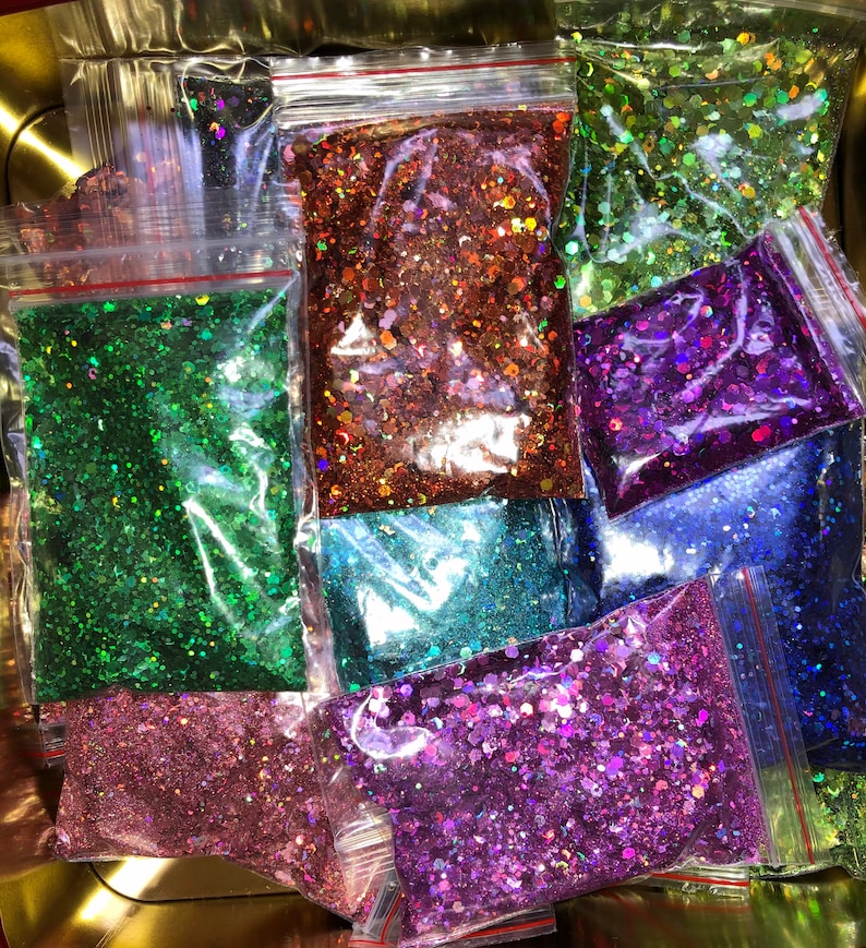 K&ouml;nnte beinhalten: Sortiment an schimmerndem Glitter in verschiedenen Farben, darunter Gr&uuml;n, Lila, Blau und Pink, verpackt in durchsichtigen Plastikt&uuml;ten mit roten Rei&szlig;verschl&uuml;ssen.