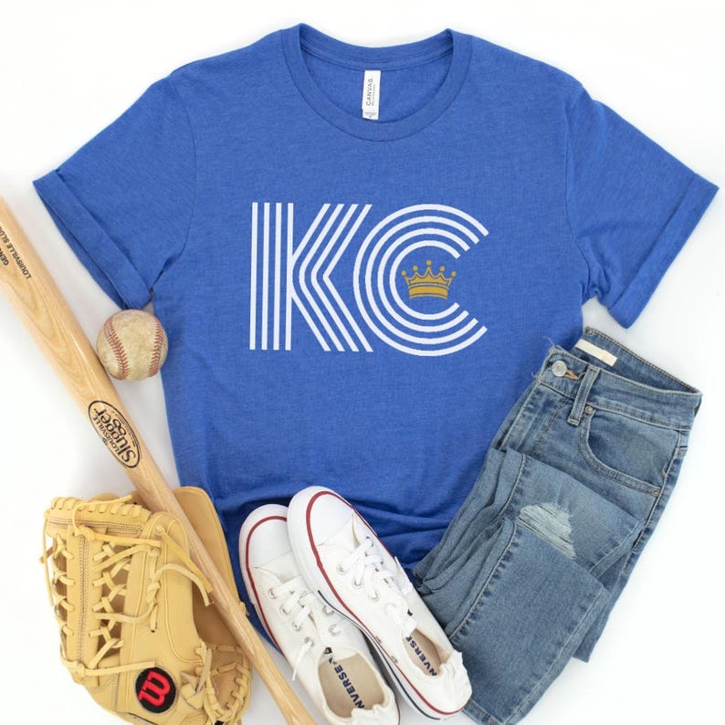 Puede incluir: Una camiseta azul real con un gr&aacute;fico blanco de las letras "KC" con una corona dorada en la parte superior. Las letras est&aacute;n rodeadas de rayas blancas.