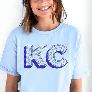 May include: A light blue short-sleeved t-shirt featuring the letters KC printed in a bold, dark blue font. The letters have a scalloped edge and are decorated with intricate, swirling line patterns, creating a distinct graphic design.