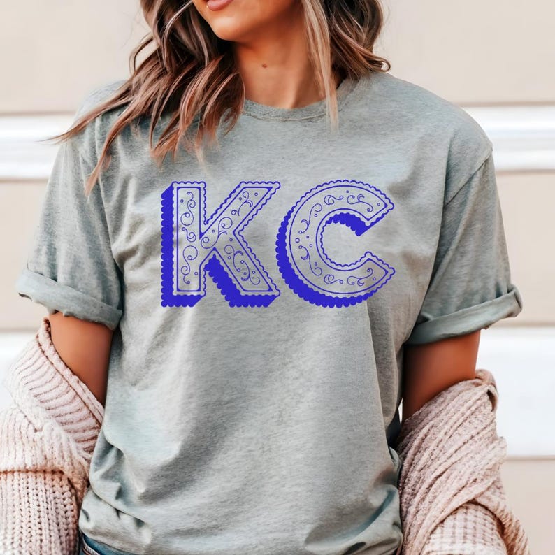 May include: A heathered sage green short-sleeve t-shirt featuring the letters KC in a bold, royal blue color. The letters have a scalloped edge and a decorative, intricate swirling line pattern inside, creating a distinct visual contrast.