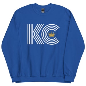 A royal blue unisex crewneck sweatshirt with a white graphic of the letters 'KC' with a gold crown over the 'C'. The letters are stylized with white lines that create a striped effect.