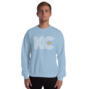 A light blue unisex crewneck sweatshirt with a white graphic of the letters 'KC' with a gold crown over the 'C'. The letters are stylized with white lines that create a striped effect.