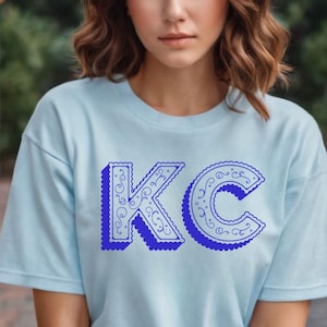 May include: A light blue short-sleeve t-shirt featuring the letters KC in a bold, dark blue font. The letters have a scalloped edge and a decorative swirling pattern inside, with a drop shadow effect that gives them a three-dimensional appearance.