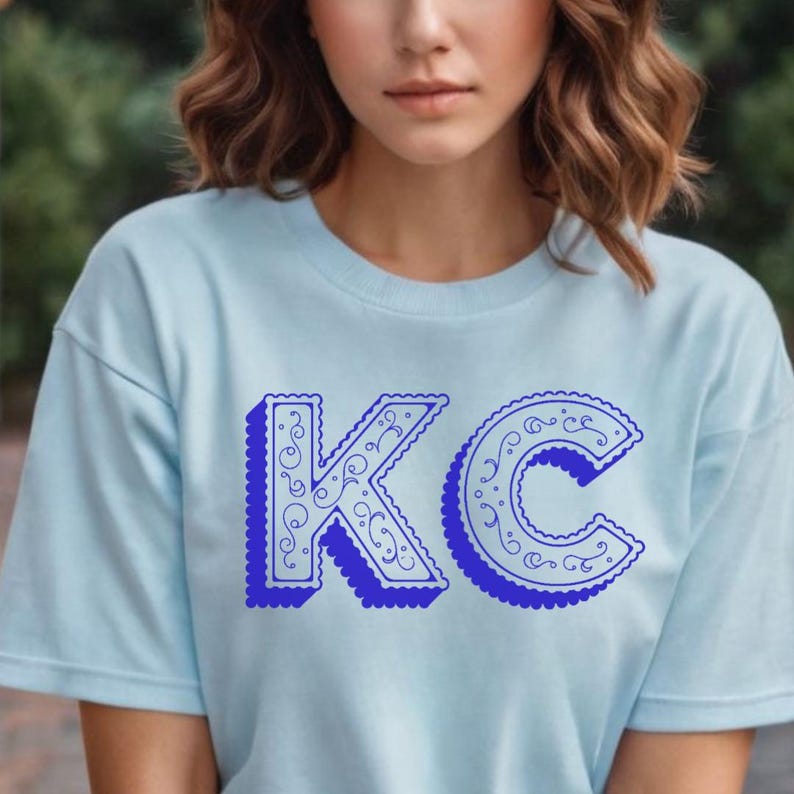 May include: A light blue short-sleeve t-shirt featuring the letters KC in a bold, dark blue font. The letters have a scalloped edge and a decorative swirling pattern inside, with a drop shadow effect that gives them a three-dimensional appearance.