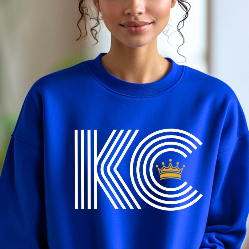 A royal blue unisex crewneck sweatshirt with a white graphic of the letters 'KC' with a gold crown over the 'C'. The letters are stylized with white lines that create a striped effect.