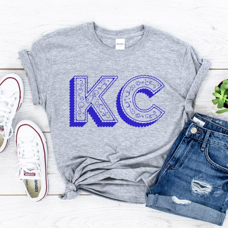 May include: A heather gray short-sleeve t-shirt featuring the letters KC in a blue, scalloped-edge font with a decorative swirl pattern inside the letters. The shirt has a crew neckline and a Gildan brand label visible at the inner collar.