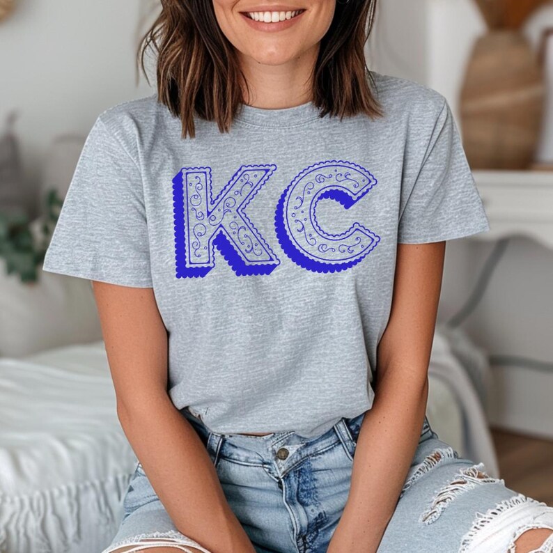 May include: A heather gray short-sleeve t-shirt featuring the letters KC in a bold blue font. The letters have a dark blue drop shadow and a decorative white filigree pattern inside the blue outlines, creating a distinct graphic design.