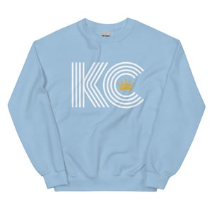 A light blue unisex crewneck sweatshirt with a white graphic of the letters 'KC' with a gold crown over the 'C'. The letters are stylized with white lines that create a striped effect.