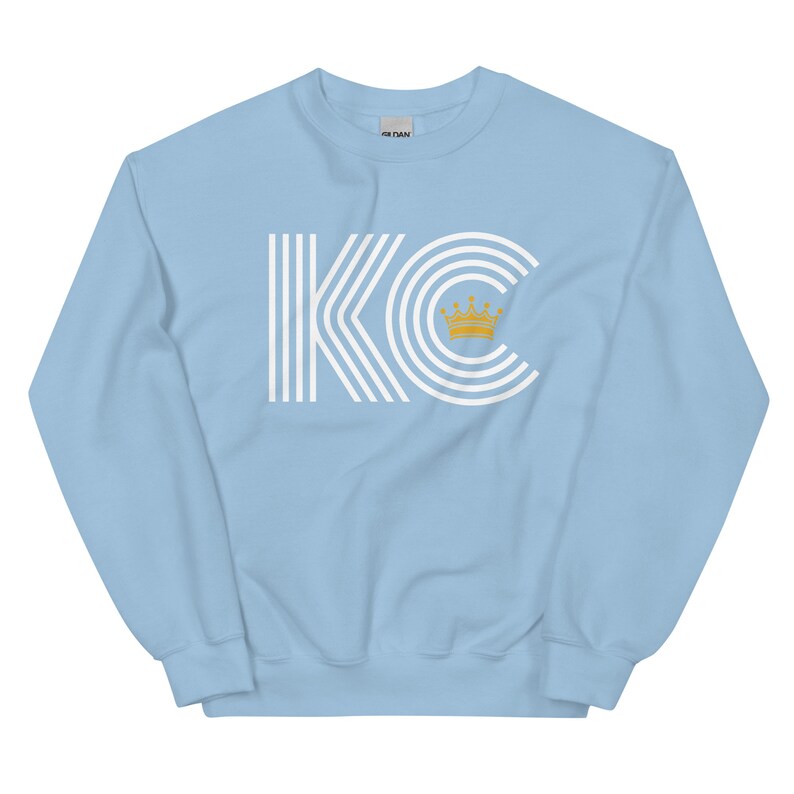A light blue unisex crewneck sweatshirt with a white graphic of the letters 'KC' with a gold crown over the 'C'. The letters are stylized with white lines that create a striped effect.