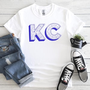 May include: A white short-sleeved t-shirt featuring the blue letters KC in a decorative, swirled font with a drop shadow effect. The shirt is displayed alongside blue denim jeans and a pair of black and white low-top canvas sneakers.