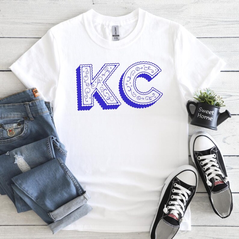 May include: A white short-sleeved t-shirt featuring the blue letters KC in a decorative, swirled font with a drop shadow effect. The shirt is displayed alongside blue denim jeans and a pair of black and white low-top canvas sneakers.