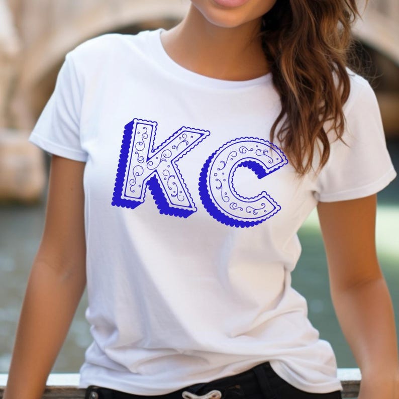 May include: A white short-sleeved t-shirt featuring the letters KC in a bold, royal blue color. The letters have a three-dimensional effect with a scalloped edge and an intricate, swirling filigree pattern printed on the front of the garment.
