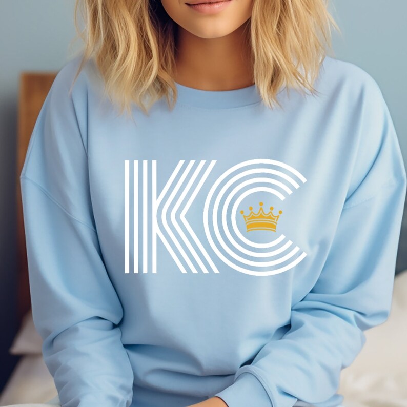 A light blue unisex crewneck sweatshirt with a white graphic of the letters 'KC' with a gold crown over the 'C'. The letters are stylized with white lines that create a striped effect.