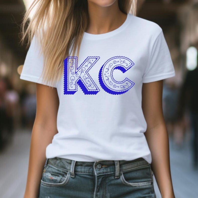 May include: A white short-sleeved t-shirt featuring the letters KC printed in a bold blue font. The letters have a decorative, intricate swirling pattern inside and a scalloped edge, creating a distinct visual effect against the plain white fabric.