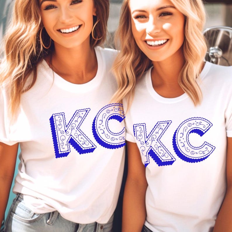 May include: A white short-sleeved t-shirt featuring the letters K and C in a bold blue font with a decorative swirl pattern inside the characters and a blue drop shadow effect. The design is printed on the front center of the garment.