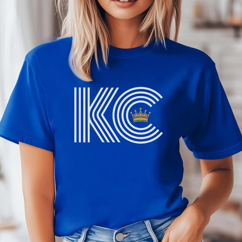 Puede incluir: Una camiseta azul real con un gr&aacute;fico blanco de las letras "KC" con una corona dorada dentro de la "C".