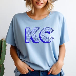 May include: A light blue short-sleeve t-shirt featuring the letters KC in a bold, dark blue font with a scalloped edge and intricate white filigree scrollwork patterns inside the letters, set against a solid light blue fabric background.