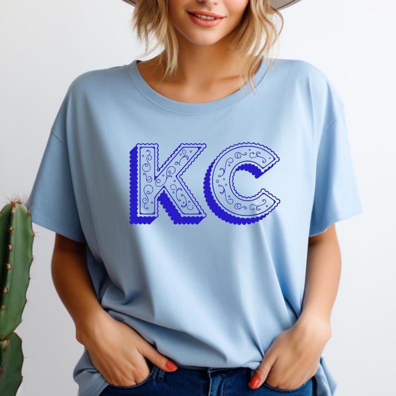May include: A light blue short-sleeve t-shirt featuring the letters KC in a bold, dark blue font with a scalloped edge and intricate white filigree scrollwork patterns inside the letters, set against a solid light blue fabric background.