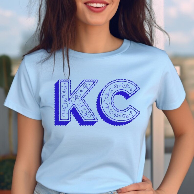 May include: A light blue short-sleeve t-shirt featuring the letters KC in a dark blue color. The letters have a scalloped edge and a decorative swirling pattern inside, with a dark blue drop shadow effect that gives them a three-dimensional look.