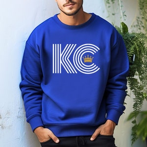 A royal blue unisex crewneck sweatshirt with a white graphic of the letters 'KC' with a gold crown over the 'C'. The letters are stylized with white lines that create a striped effect.