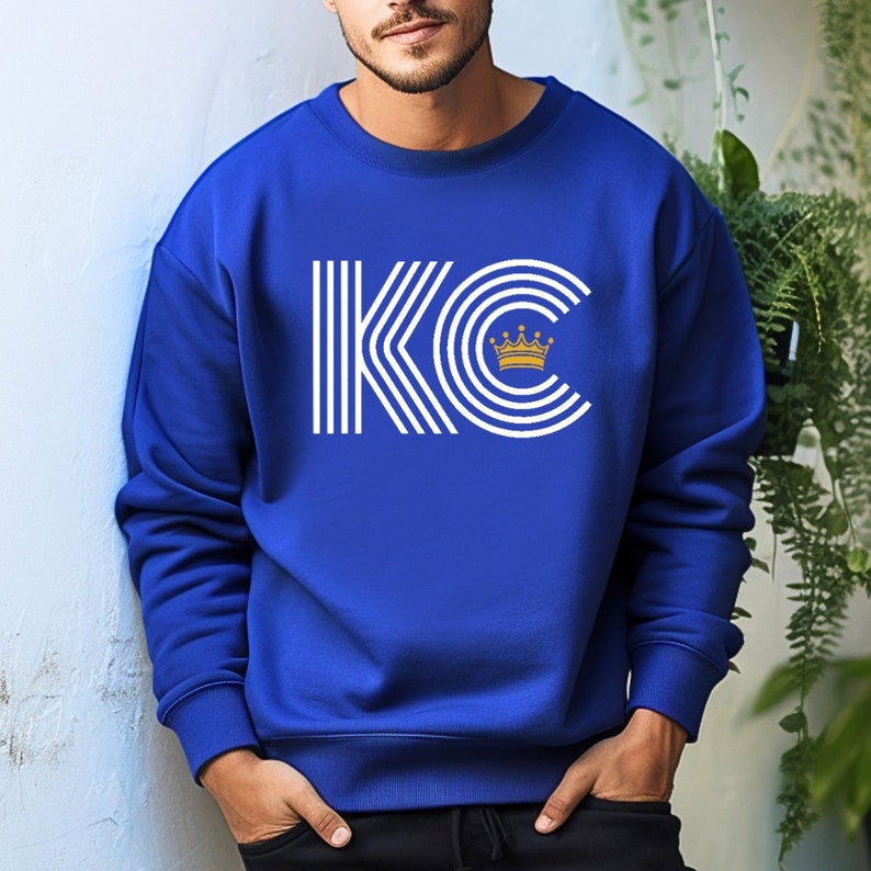 A royal blue unisex crewneck sweatshirt with a white graphic of the letters 'KC' with a gold crown over the 'C'. The letters are stylized with white lines that create a striped effect.