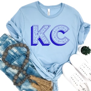 May include: A light blue crew neck t-shirt featuring the letters KC in a dark blue, ornate, swirled font with a scalloped edge and a drop shadow effect. The shirt is displayed with blue tie-dye fabric, a string of grey wooden beads, and white sneakers.