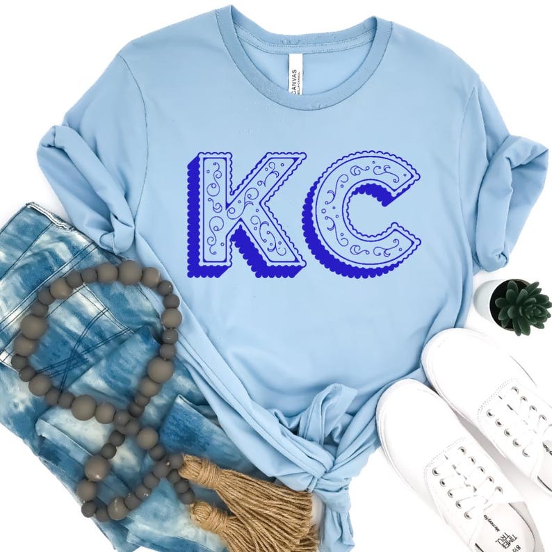 May include: A light blue crew neck t-shirt featuring the letters KC in a dark blue, ornate, swirled font with a scalloped edge and a drop shadow effect. The shirt is displayed with blue tie-dye fabric, a string of grey wooden beads, and white sneakers.