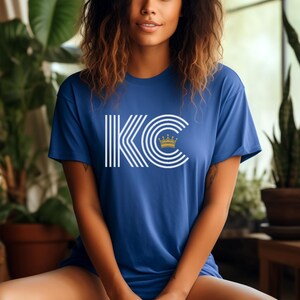 Puede incluir: Una camiseta azul real con un gr&aacute;fico blanco de las letras "KC" con una corona dorada en la parte superior. Las letras est&aacute;n rodeadas de rayas blancas.