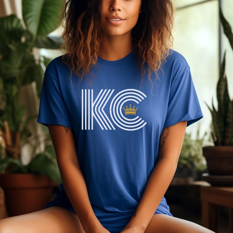 Puede incluir: Una camiseta azul real con un gr&aacute;fico blanco de las letras "KC" con una corona dorada en la parte superior. Las letras est&aacute;n rodeadas de rayas blancas.