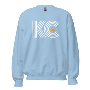 A light blue unisex crewneck sweatshirt with a white graphic of the letters 'KC' with a gold crown over the 'C'. The letters are stylized with white lines that create a striped effect.