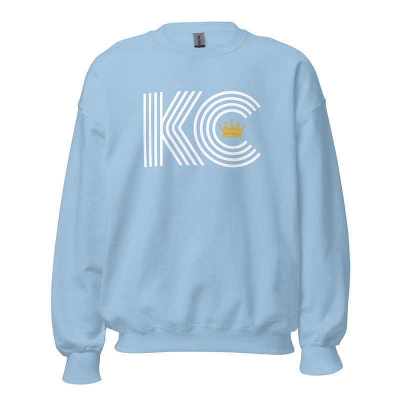 A light blue unisex crewneck sweatshirt with a white graphic of the letters 'KC' with a gold crown over the 'C'. The letters are stylized with white lines that create a striped effect.
