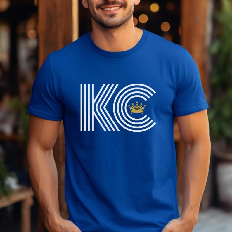 Puede incluir: Una camiseta azul real con un gr&aacute;fico blanco de las letras "KC" con una corona dorada sobre la "C".