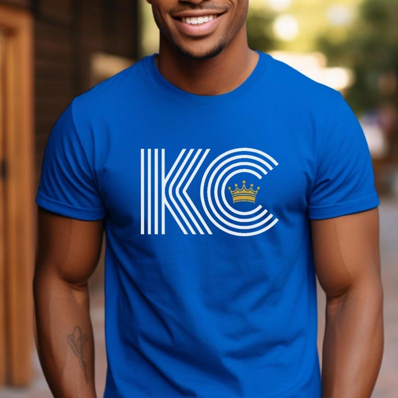 Puede incluir: Una camiseta azul real con un gr&aacute;fico blanco de las letras "KC" con una corona dorada sobre la "C".