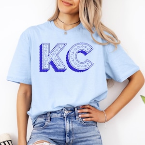May include: A light blue short-sleeve t-shirt featuring the letters KC in a bold, royal blue font. The letters have a scalloped edge and a white interior filled with intricate, swirling line patterns, creating a decorative graphic design.