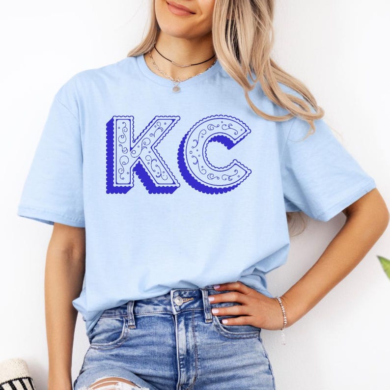 May include: A light blue short-sleeve t-shirt featuring the letters KC in a bold, royal blue font. The letters have a scalloped edge and a white interior filled with intricate, swirling line patterns, creating a decorative graphic design.