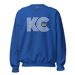 A royal blue unisex crewneck sweatshirt with a white graphic of the letters 'KC' with a gold crown over the 'C'. The letters are stylized with white lines that create a striped effect.
