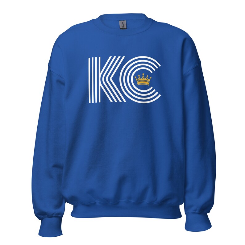A royal blue unisex crewneck sweatshirt with a white graphic of the letters 'KC' with a gold crown over the 'C'. The letters are stylized with white lines that create a striped effect.