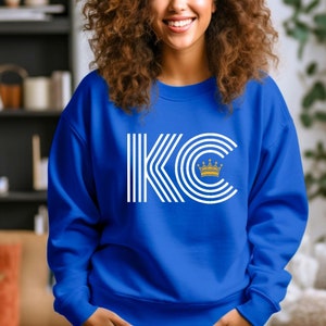 A royal blue unisex crewneck sweatshirt with a white graphic of the letters 'KC' with a gold crown over the 'C'. The letters are stylized with white lines that create a striped effect.