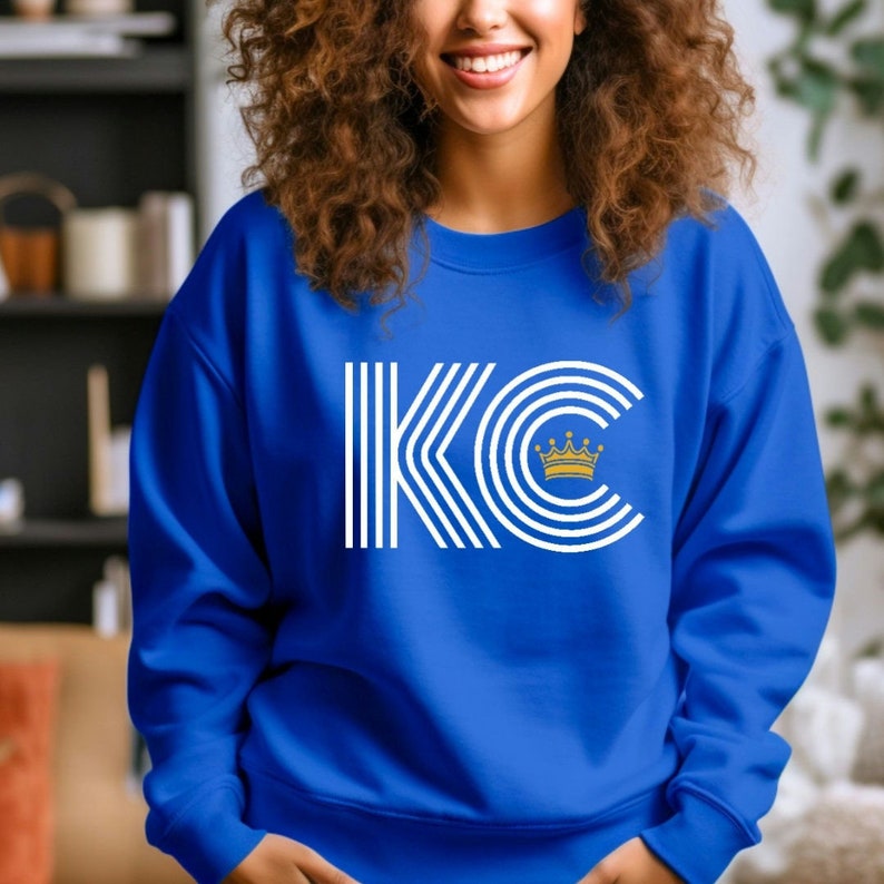 A royal blue unisex crewneck sweatshirt with a white graphic of the letters 'KC' with a gold crown over the 'C'. The letters are stylized with white lines that create a striped effect.