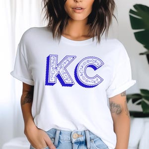 May include: A white short-sleeved t-shirt featuring the letters KC printed in a bold blue color. The letters have a decorative, swirling filigree pattern inside and a scalloped edge outline, creating a distinct graphic design on the front.
