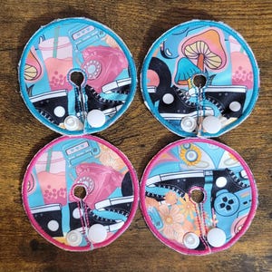 Peut inclure: Quatre coussinets d'allaitement ronds en tissu avec des motifs colorés. Chaque coussinet présente une basket noire, un téléphone rotatif et d'autres graphiques rétro sur un fond turquoise. Les coussinets ont une fermeture à bouton-pression blanche et sont bordés de tissu bleu ou rose.