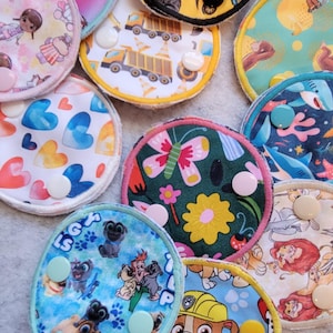May include: A collection of colorful, round fabric pads with various cartoon and nature-themed designs. The pads feature images of trucks, butterflies, hearts, and cartoon characters. Each pad has a button closure.