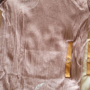 May include: A close-up of several baby bodysuits in shades of brown and yellow. The brown bodysuit has a ribbed texture and a decorative logo. The yellow bodysuit is visible in the background. The bodysuits have snap closures and the Gerber brand label.