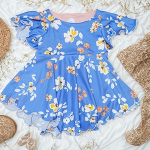 May include: A blue dress with a floral pattern featuring white and yellow flowers, and peach accents. The dress has ruffled sleeves and a scalloped hem. A straw hat, patterned fabric, and sandals are also visible.